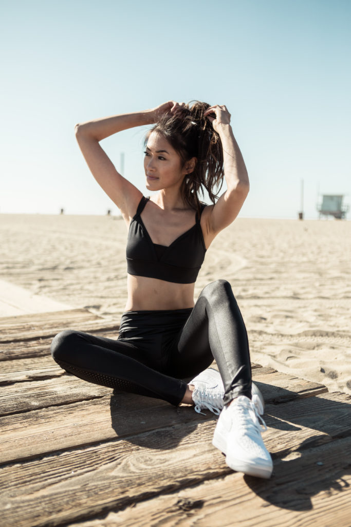 woman at the beach and wearing all black sportswear, white running shoes, and tying her hair 